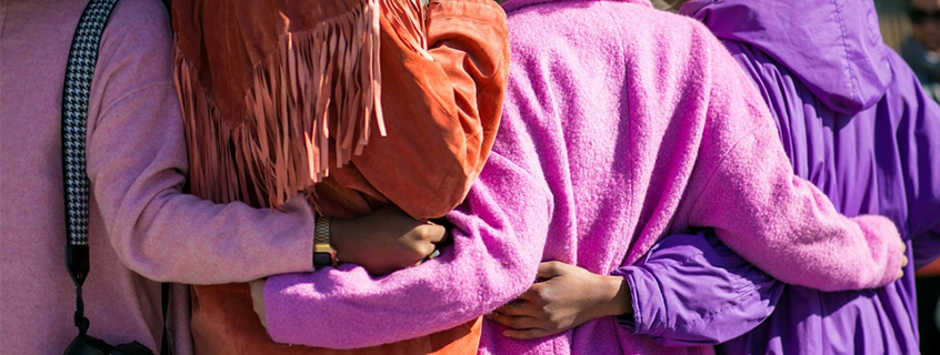 Four girls in brightly colored pink, purple, and orange jackets have their arms around each other. Photo is from behind